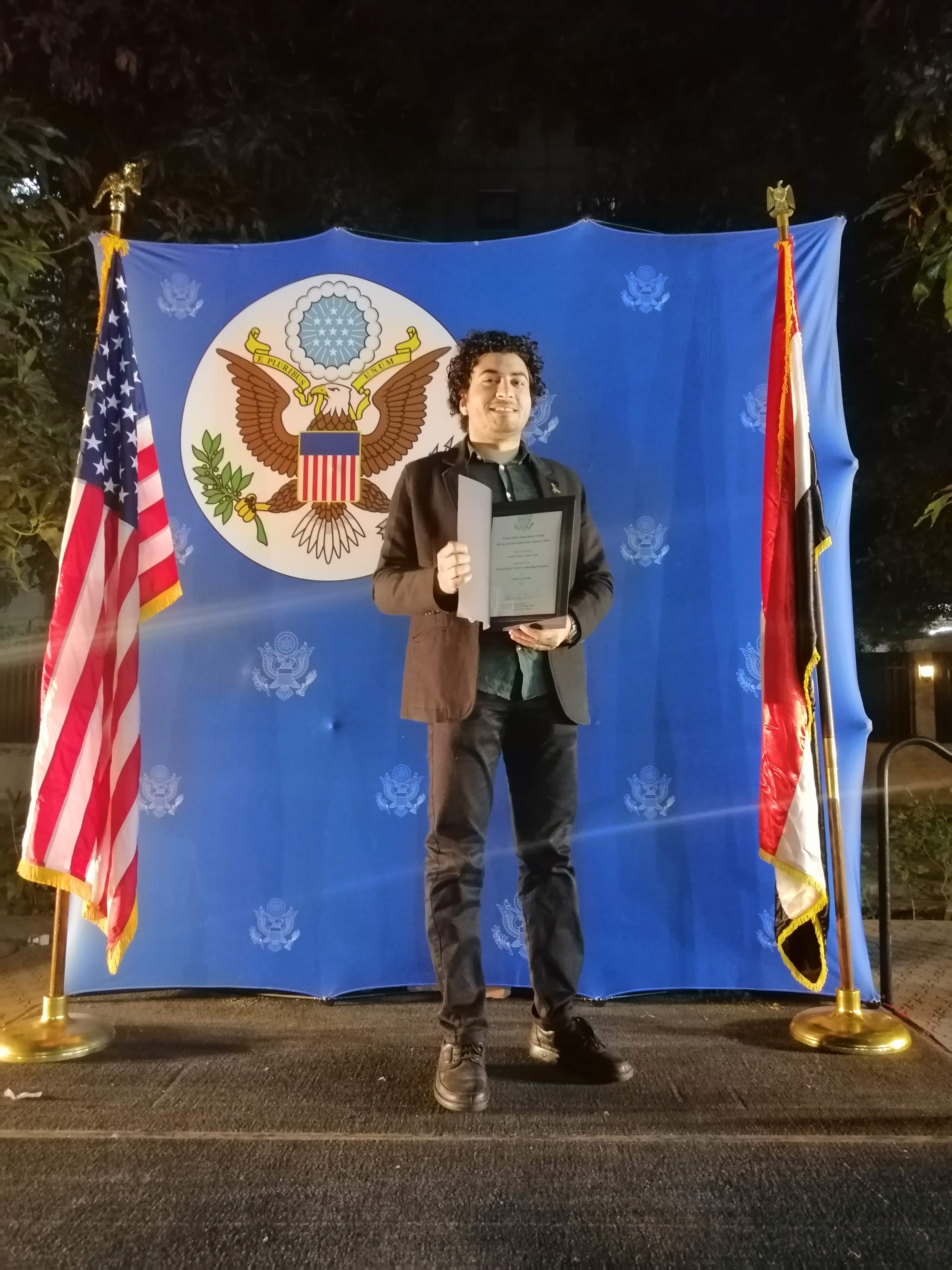 Person holding a certificate in front of U.S. and Egyptian flags during an award ceremony.
