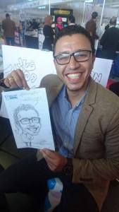Man smiling and holding a caricature drawing of himself at an event, inviting others to take a picture.