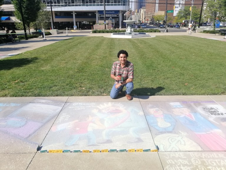 Man kneeling beside chalk drawings on a sidewalk in a park.