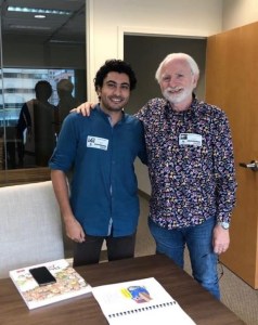 Cartoonists Sameh Samir and KAL smiling and interacting during an office meeting with documents on the table.