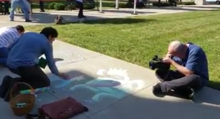 People drawing with chalk on a sidewalk while being filmed by a cameraman during a public art event.