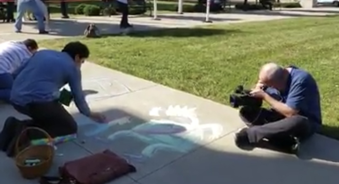 People drawing with chalk on a sidewalk while being filmed by a cameraman during a public art event.
