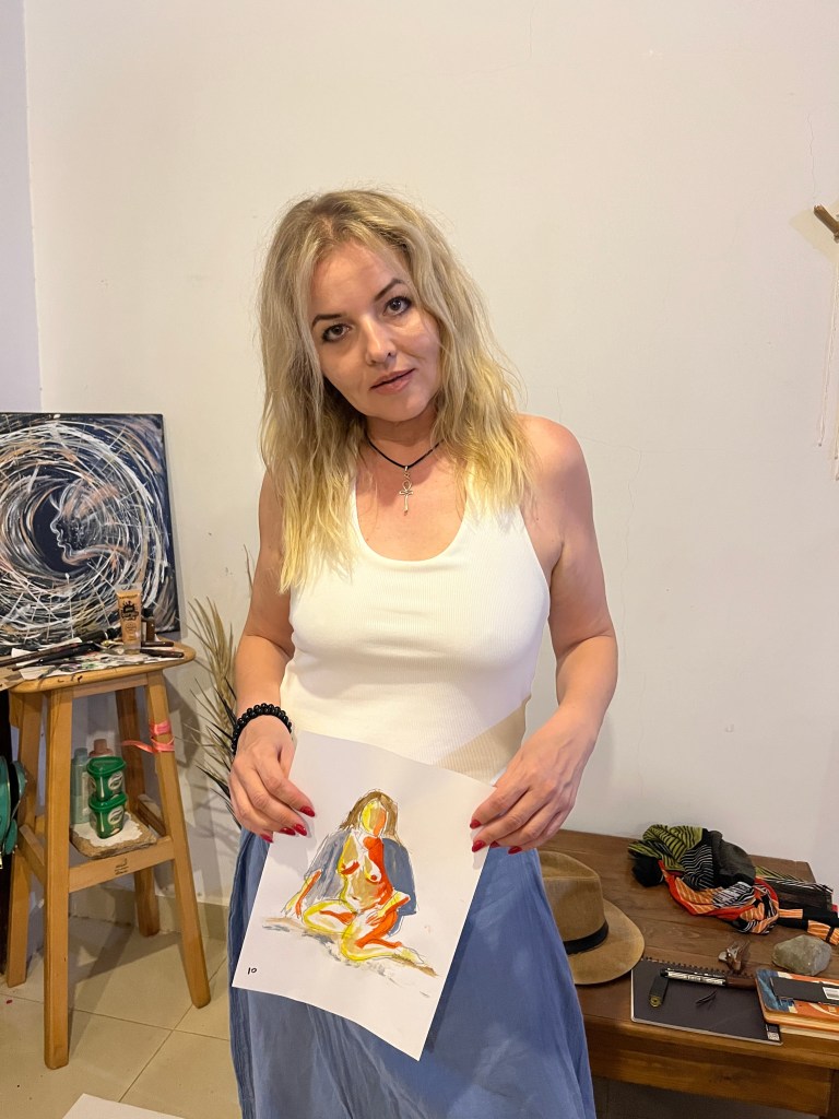 Woman holding a painting in an art studio, surrounded by art supplies and canvases.